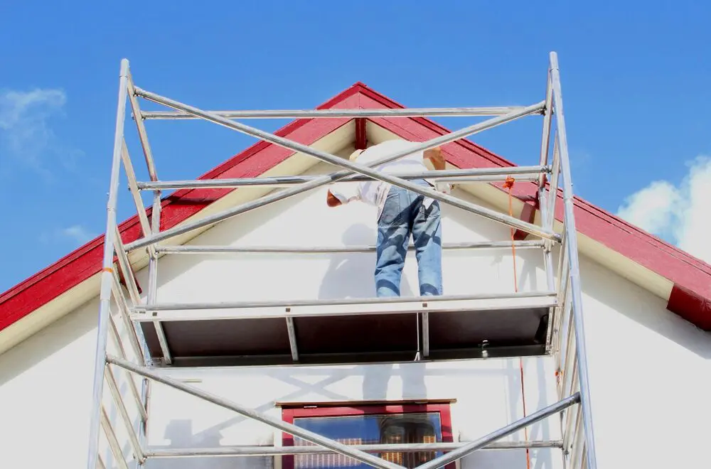 A pro painter standing on scaffolding, painting a house’s exterior trim under a bright blue sky.