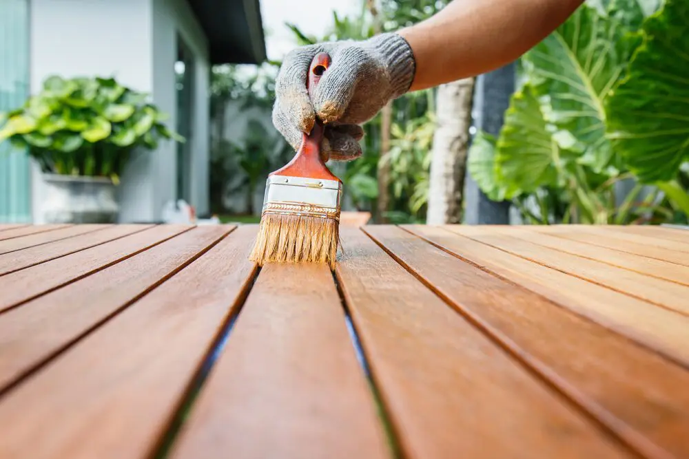 Gloved hand applies stain with brush on wooden surface, showing proper wood deck staining technique for longevity.