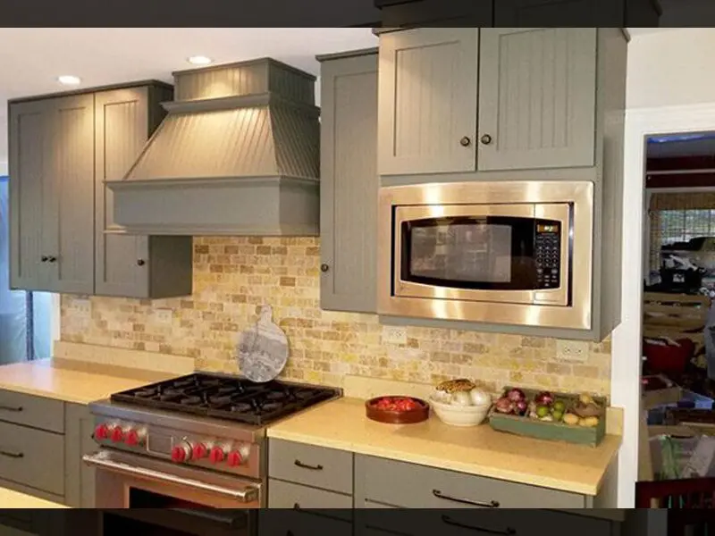 Gray stained wood cabinets with stainless microwave and hood vent in modern kitchen with light stone backsplash.