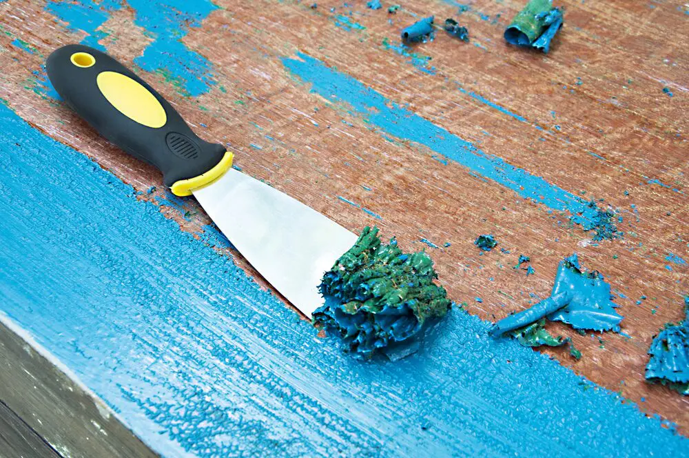 A close-up of a putty knife scraping blue peeling paint from a wooden surface, showing preparation to repaint and prevent paint from fading due to weather exposure.