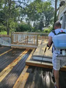 A worker using a backpack sprayer to clean and treat a wooden deck with dark-stained and newly replaced boards.