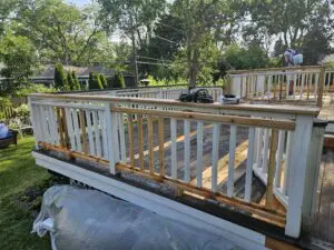 A deck restoration project in progress, with partially stripped railings and darkened wood being prepped for staining by a worker using a sprayer.