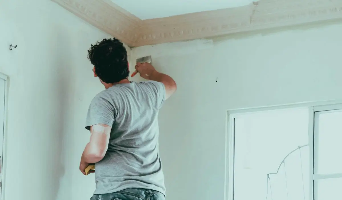 A person painting an interior wall using a paintbrush, focusing on the edges near the ceiling with natural light coming through a nearby window.