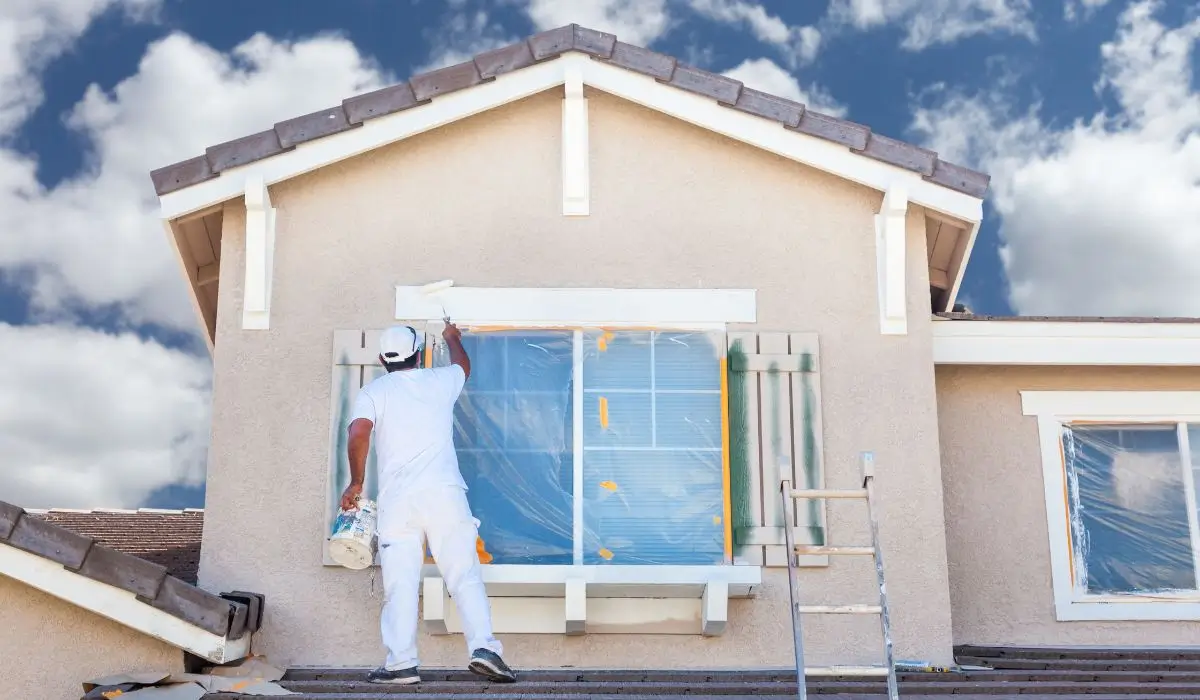 A pro painter in white overalls painting the exterior of a house using a roller and ladder.