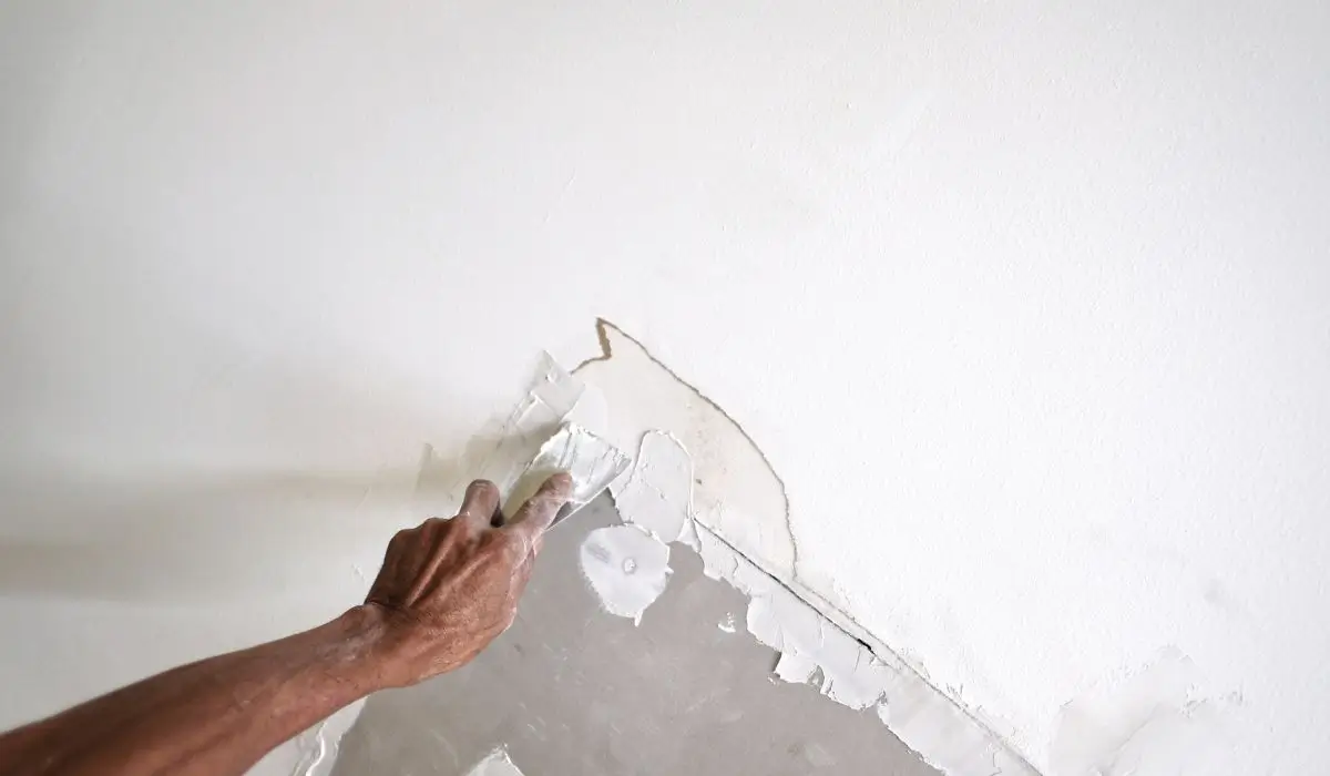 A worker carefully scraping peeling paint and applying patching compound to repair drywall and fix wall damage.