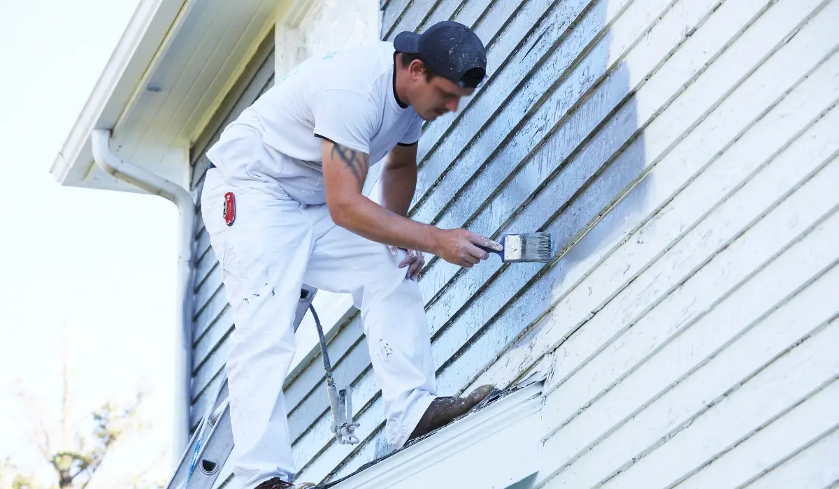 A professional painter in fall gear applying exterior paint to weathered siding on a two-story home in Barrington South, preparing the surface before winter conditions arrive.